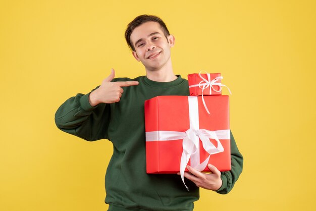 노란색 배경에 서 크리스마스 선물을 가리키는 크리스마스 선물 전면보기 젊은 남자