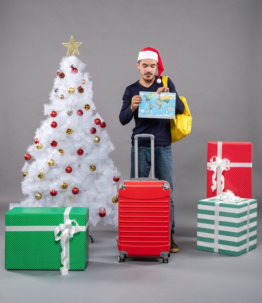 Вид спереди молодой человек с желтым рюкзаком, держащий и смотрящий на карту возле рождественской елки и подарков на сером изолированном