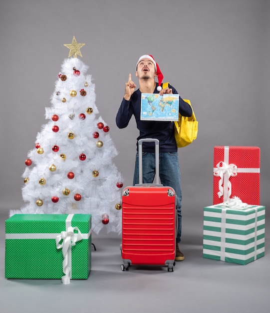 Front view young man with yellow backpack holding map looking up near white xmas tree on grey isolated