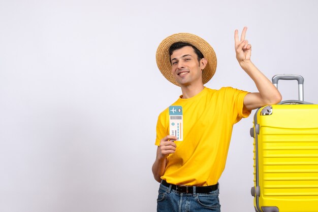 Front view young man in yellow t-shirt standing near yellow suitcase holding ticket making v sign