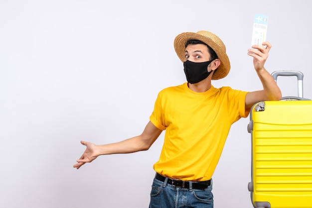 Front view young man in yellow t-shirt standing near yellow suitcase holding up airplane ticket