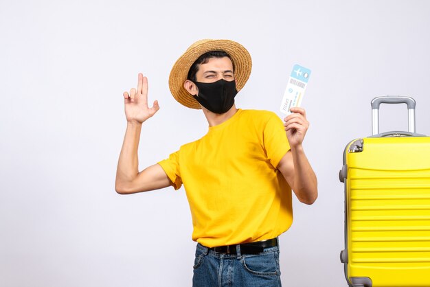 Front view young man in yellow t-shirt standing near yellow suitcase holding up travel ticket making finger gun