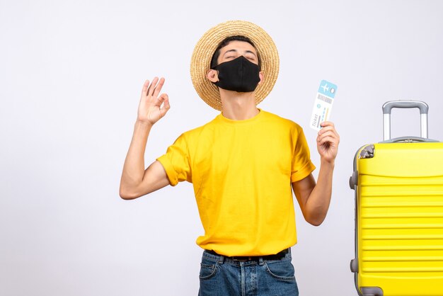 黄色のTシャツを着た若い男が黄色いスーツケースの近くに立って旅行チケットを持ってOKサインを作る正面図