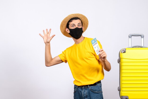 Front view young man in yellow t-shirt standing near yellow suitcase holding up travel ticket waving hand