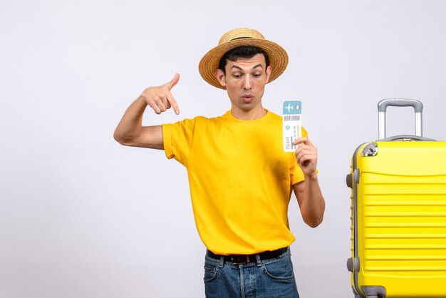 Front view young man in yellow t-shirt standing near yellow suitcase pointing at below