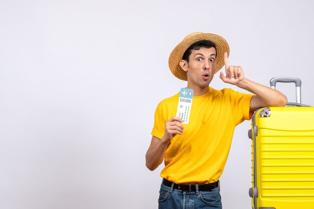 Front view young man in yellow t-shirt standing near yellow suitcase surprising with an idea