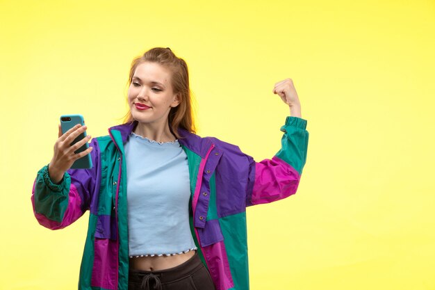 A front view young modern woman in blue shirt black trousers colorful jacket smiling posing using phone flexing