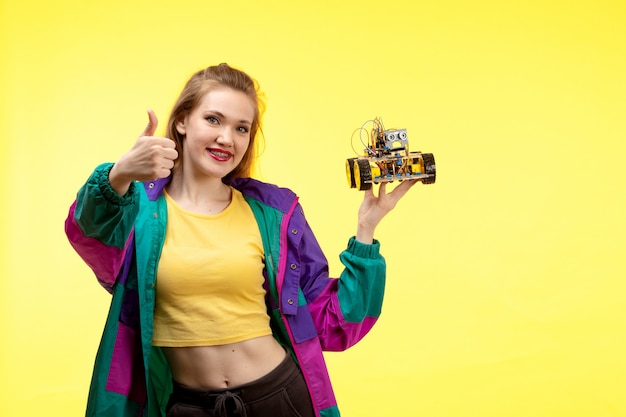 黄色のシャツの黒いズボンと幸せな表情をポーズのおもちゃの車を保持しているカラフルなジャケットの正面の若い現代女性