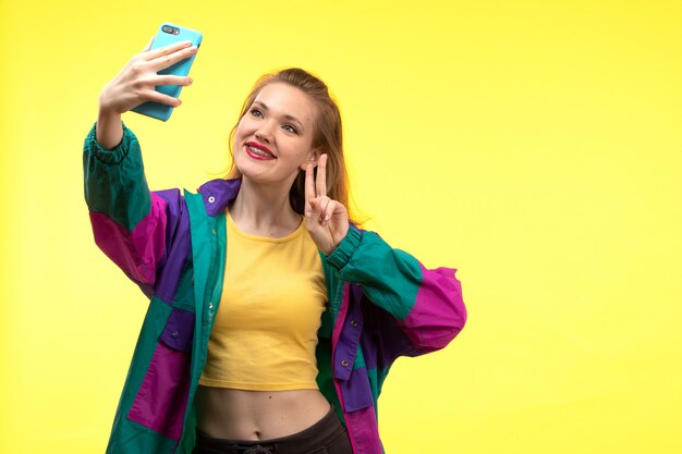 黄色いシャツの黒のズボンと携帯電話の幸せな表情でselfieを取ってカラフルなジャケットの正面の若い現代女性