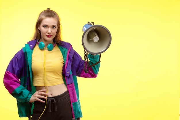 Free photo a front view young modern woman in yellow shirt black trousers and colorful jacket with colored earphones holding megaphone posing