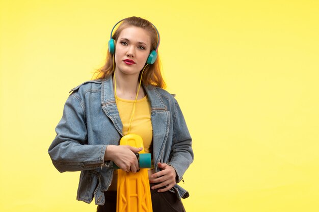 A front view young modern woman in yellow shirt black trousers and jean coat holding skateboard with colored earphones