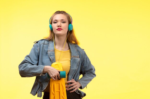 A front view young modern woman in yellow shirt black trousers and jean coat holding skateboard with colored earphones