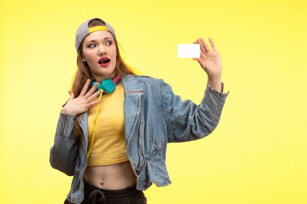 A front view young modern woman in yellow shirt black trousers and jean coat with colored earphones holding white card posing