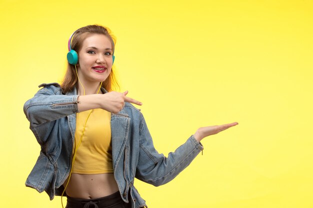 A front view young modern woman in yellow shirt black trousers and jean coat with colored earphones listening to music posing smiling