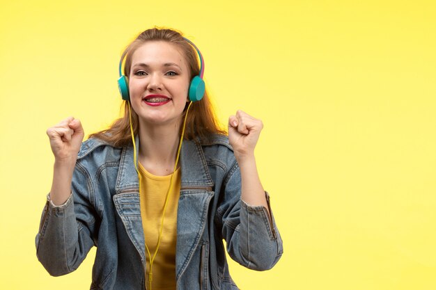 A front view young modern woman in yellow shirt black trousers and jean coat with colored earphones listening to music posing