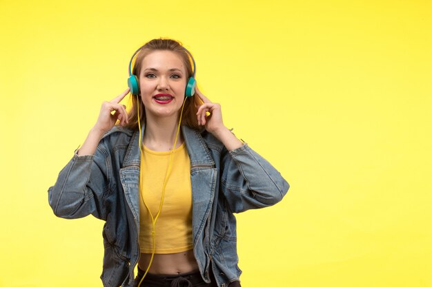 A front view young modern woman in yellow shirt black trousers and jean coat with colored earphones listening to music posing