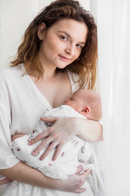 Вид спереди молодой матери с ребенком, стоя возле белой занавеской