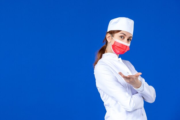 Вид спереди молодая медсестра в медицинском костюме с красной маской на синей стене