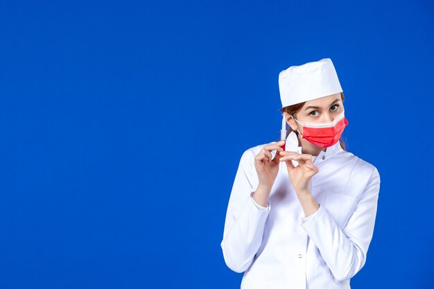 Front view young nurse in medical suit with red mask and injection in her hands on blue wall