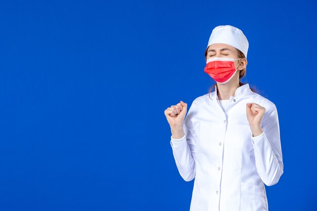 Front view young nurse in medical suit with red protective mask on blue wall