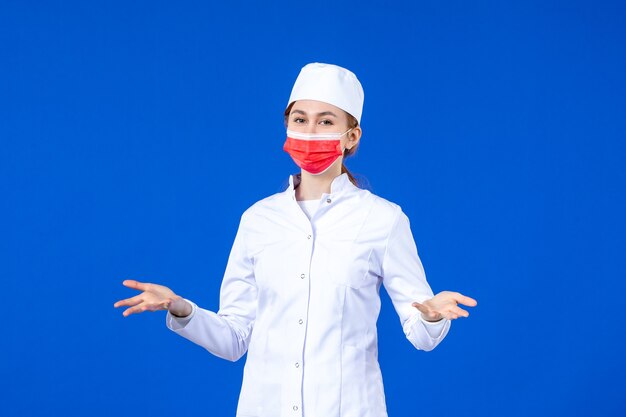 Giovane infermiera di vista frontale in vestito medico con maschera protettiva rossa sulla parete blu