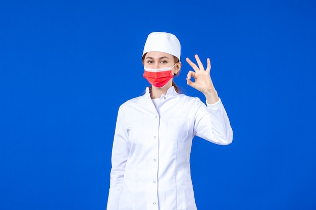 Front view young nurse in medical suit with red protective mask on blue wall