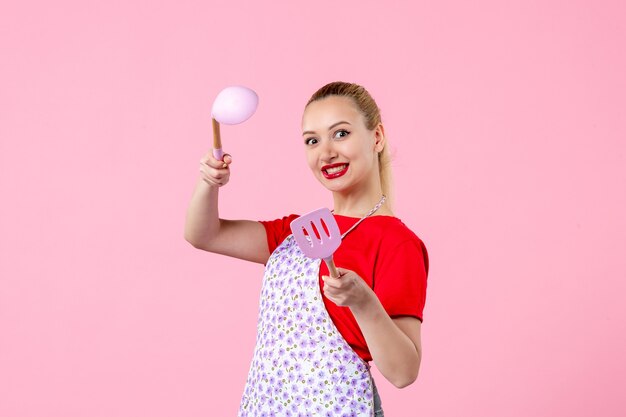 Front view young pretty housewife in cape holding spoons on pink wall