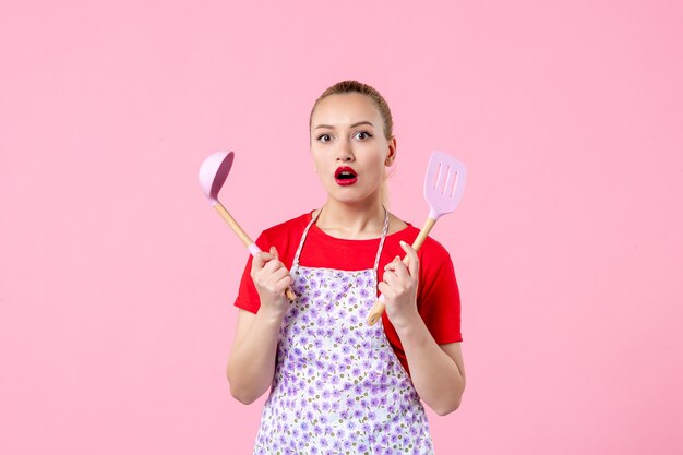 Front view young pretty housewife in cape holding spoons on pink wall