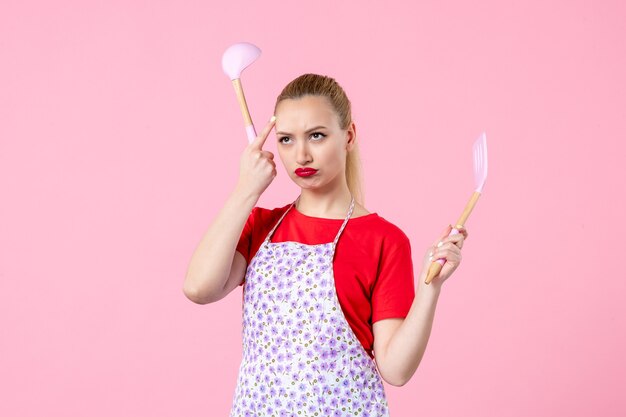 Front view young pretty housewife in cape holding spoons on pink wall