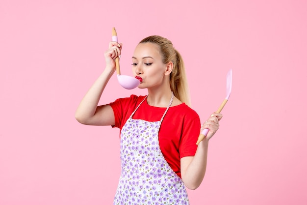 Front view young pretty housewife in cape tasting something from spoon on pink wall