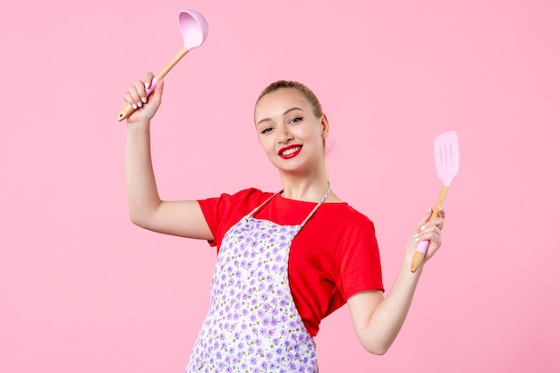 Бесплатное фото Вид спереди молодая красивая домохозяйка в накидке с ложками на розовой стене