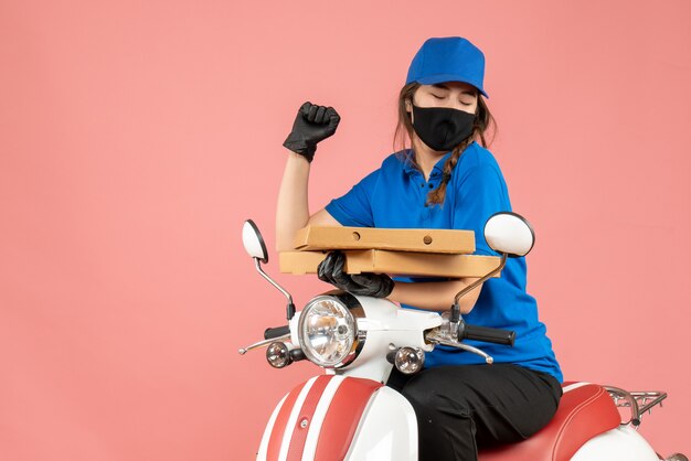 Front view of young proud female courier wearing medical mask and gloves sitting on scooter delivering orders on pastel peach background