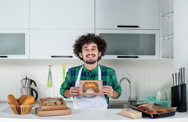Front view of young proud man holding freshly-baked pastry in a small box in the white kitchen