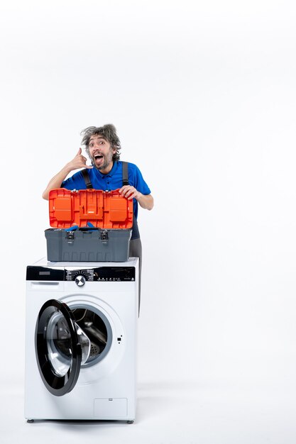 Front view of young repairman making call me sign behind washing machine on white wall