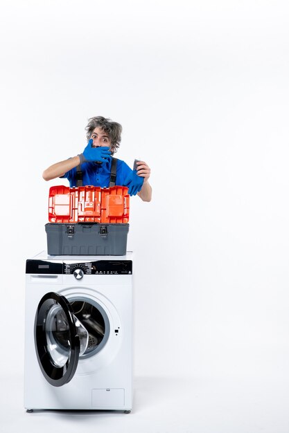 Front view of young repairman putting hand to his mouth behind washing machine on white wall