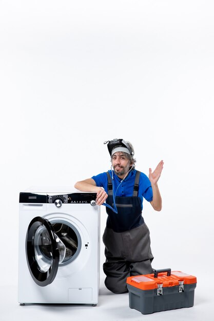 Front view of young repairman sitting near washer raising his hand on white wall