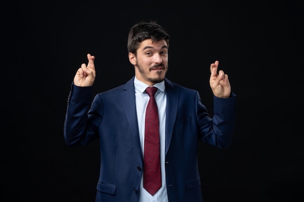 Front view of young and satisfied bearded man crossing his fingers on isolated dark wall