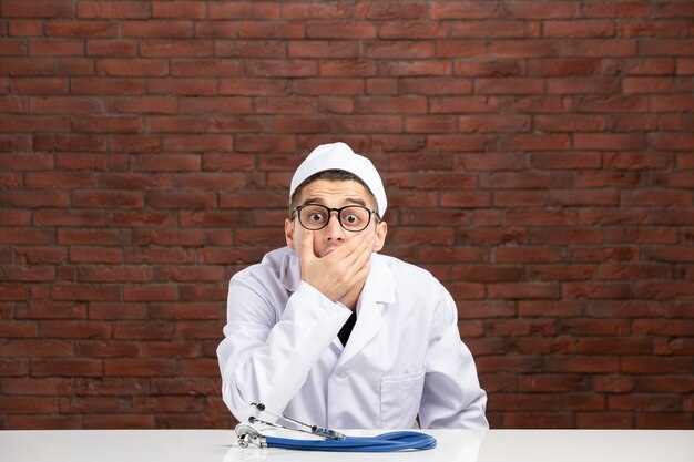 Front view young shocked doctor in white medical suit on brown bricks wall