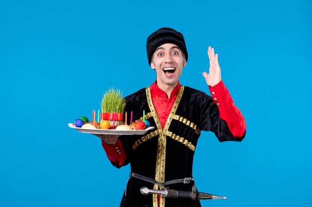 Front view of young shocked man in traditional dress holding tray filled with national confectionery on blue background