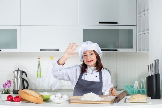 Front view of young smiling female chef in uniform saying hello to someone in the white kitchen