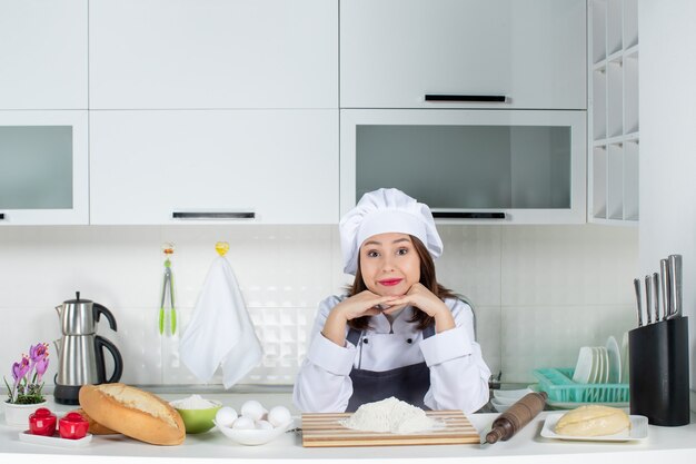 Вид спереди молодой улыбающейся женщины-шеф-повара в униформе, стоящей за столом с продуктами на разделочной доске, положив руки под подбородок на белой кухне