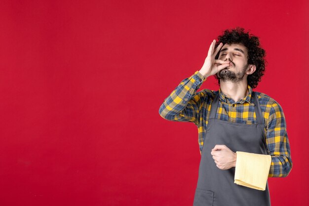 Front view of of young smiling male server with curly hair holding towel making perfect gesture standing on isolated red wall