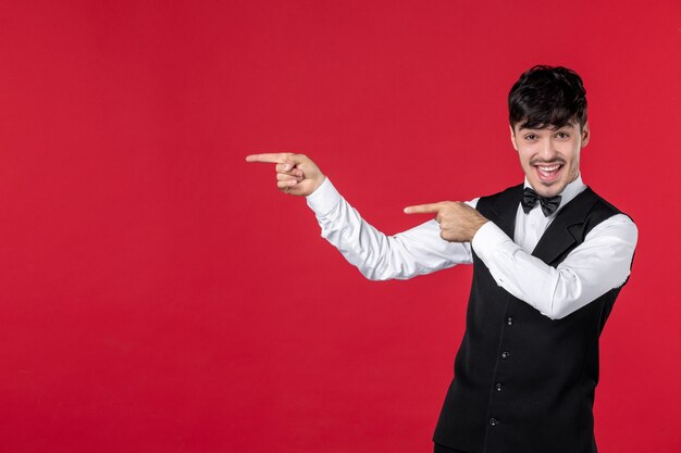 Front view of of young smiling male waiter in a uniform with bowtie on neck and pointing up on the right side with both hands on red wall