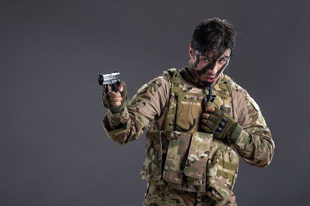Вид спереди молодого солдата в камуфляже, направленного из пистолета на темную стену