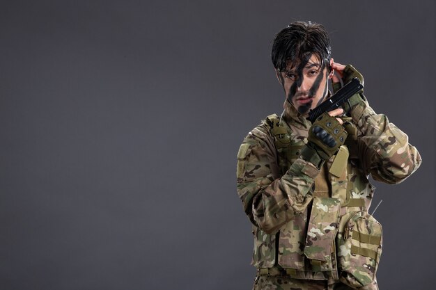 Вид спереди молодого солдата в камуфляже с пистолетом на темной стене