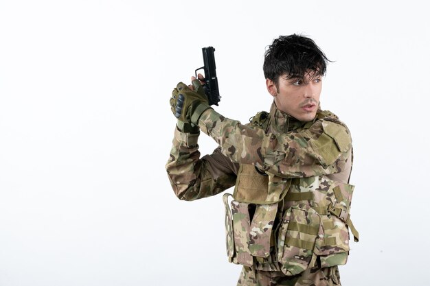 Вид спереди молодого солдата в камуфляже с пушкой на белой стене