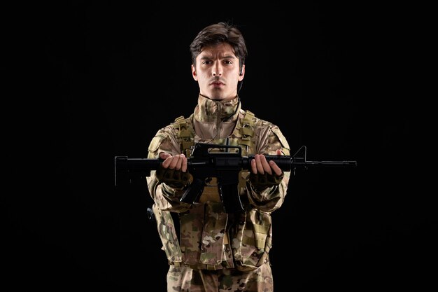 Front view of young soldier in uniform holding rifle on black wall