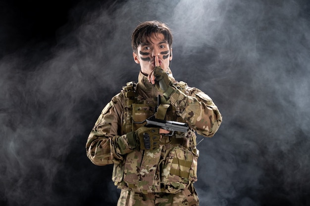 Front view of young soldier in uniform with gun black wall