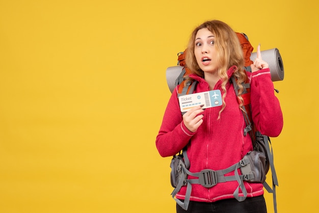 Front view of young surprised travelling girl in medical mask holding ticket and pointing up