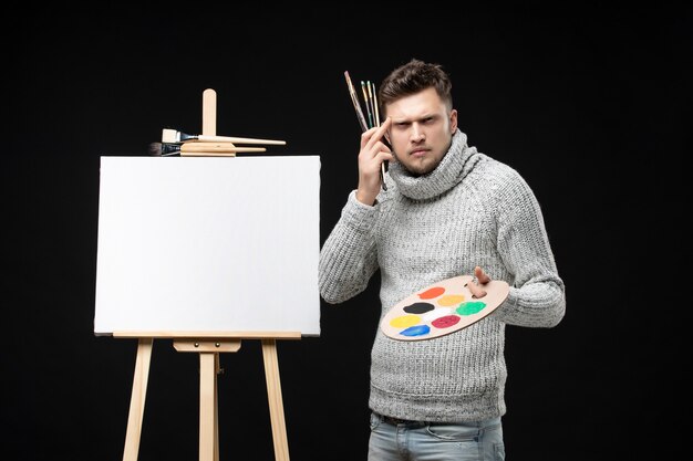 Вид спереди молодого талантливого сбитого с толку художника-мужчину, держащего картину маслом смешанных цветов на палитре и кисти на черном